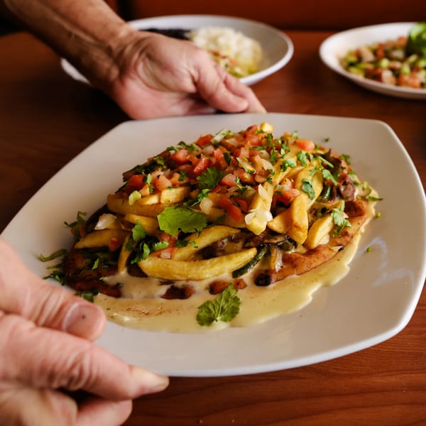 Hands serving a plate of Mexican fries topped with pico de gallo and melted cheese.