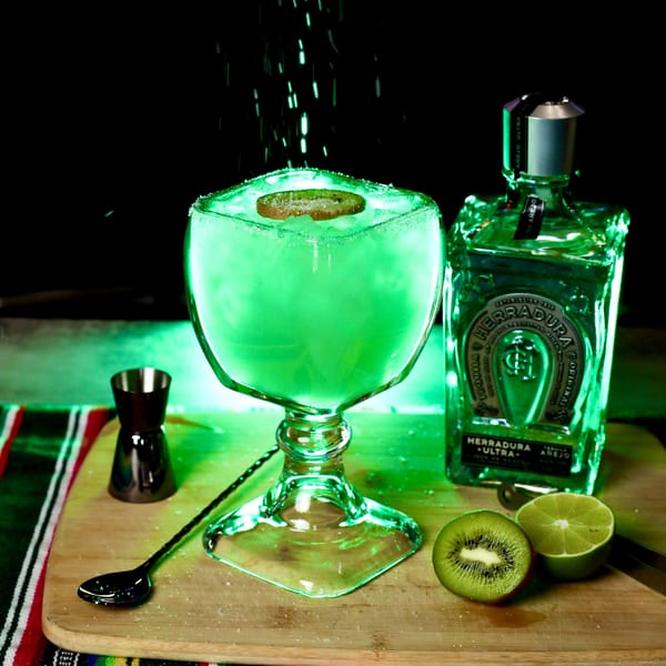 A glowing green margarita with a kiwi slice, tequila bottle, and lime on a wooden board.