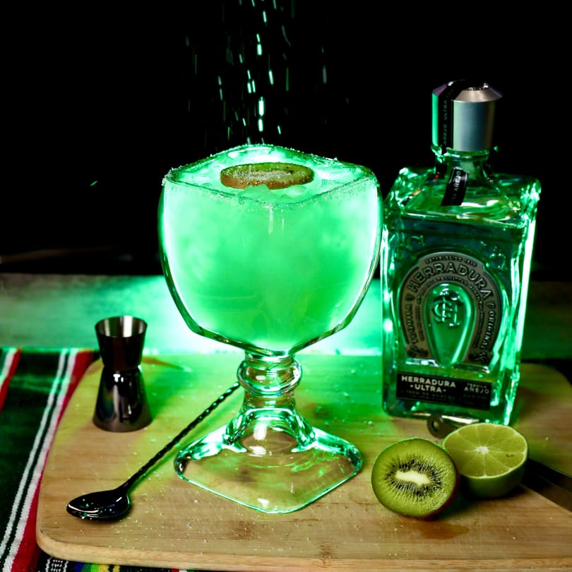 A glowing green margarita with a kiwi slice, tequila bottle, and lime on a wooden board.
