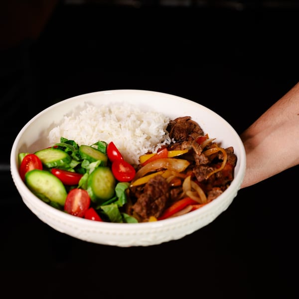 A hand holding a white bowl filled with rice, steak fajitas, and a fresh salad.