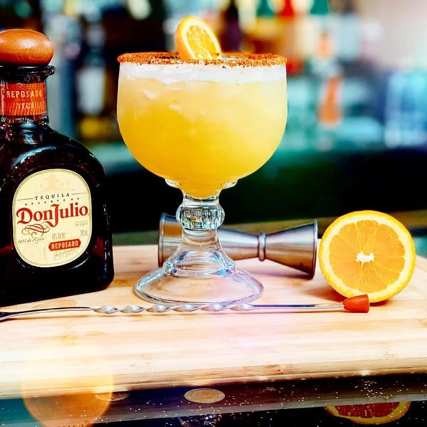 A Don Julio Reposado tequila bottle next to a margarita garnished with an orange slice and chili salt.