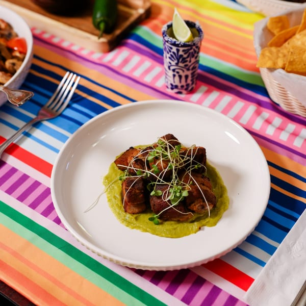 Mexican dish with cubed meat on green sauce, garnished with sprouts, on a colorful striped tablecloth.