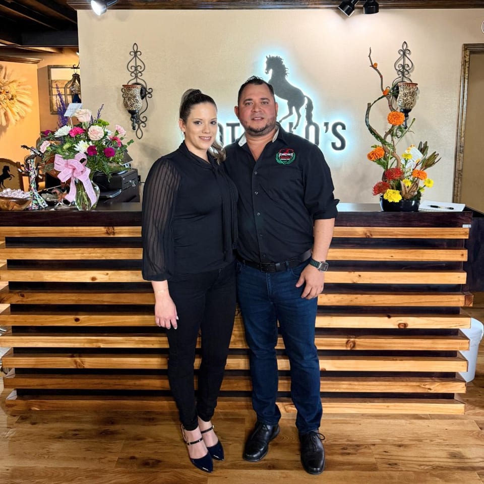 A man and woman stand in front of a Potrillo's Mexican Grill sign with a horse logo.