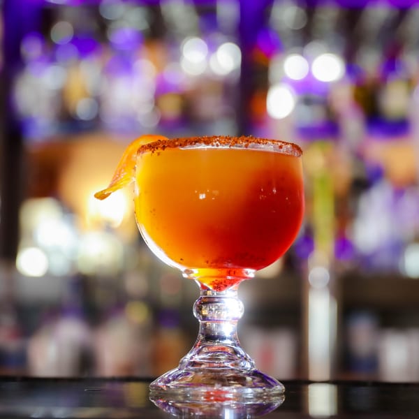 A vibrant red and orange Mexican cocktail with a chili-lime rim and orange slice, on a bar.