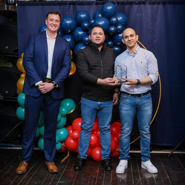 Three men posing with an award in front of a blue curtain and colorful balloons.