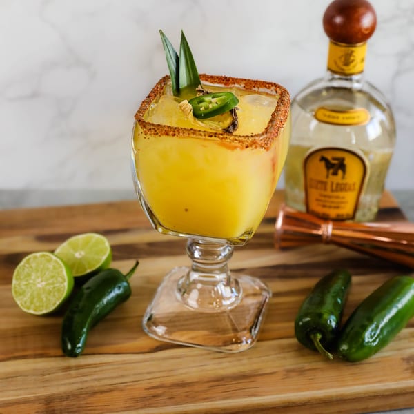 Spicy mango margarita with chili-lime rim, garnished with jalapeño and pineapple leaves, on a wooden board.