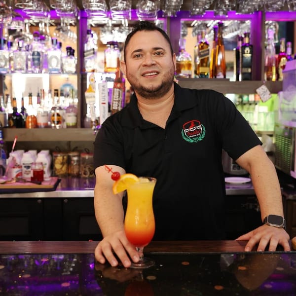A smiling bartender in a black Potrillo's Mexican Grill shirt presents a colorful cocktail.
