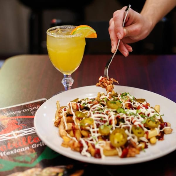 A hand forks a piece of chicken from loaded fries with jalapeños and sauces, next to a margarita.