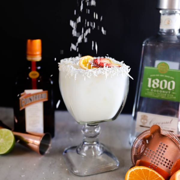 Coconut margarita with fruit garnishes and falling coconut flakes, with liquor bottles in the background.