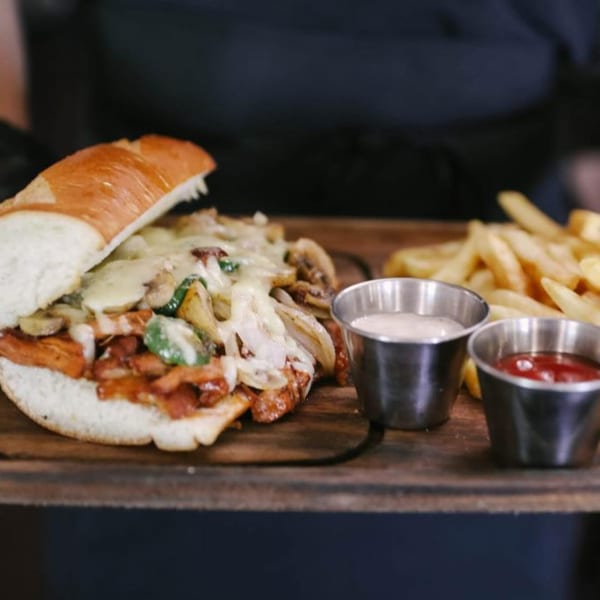 Cheesy steak and mushroom sandwich with fries, ketchup, and ranch on a wooden board.