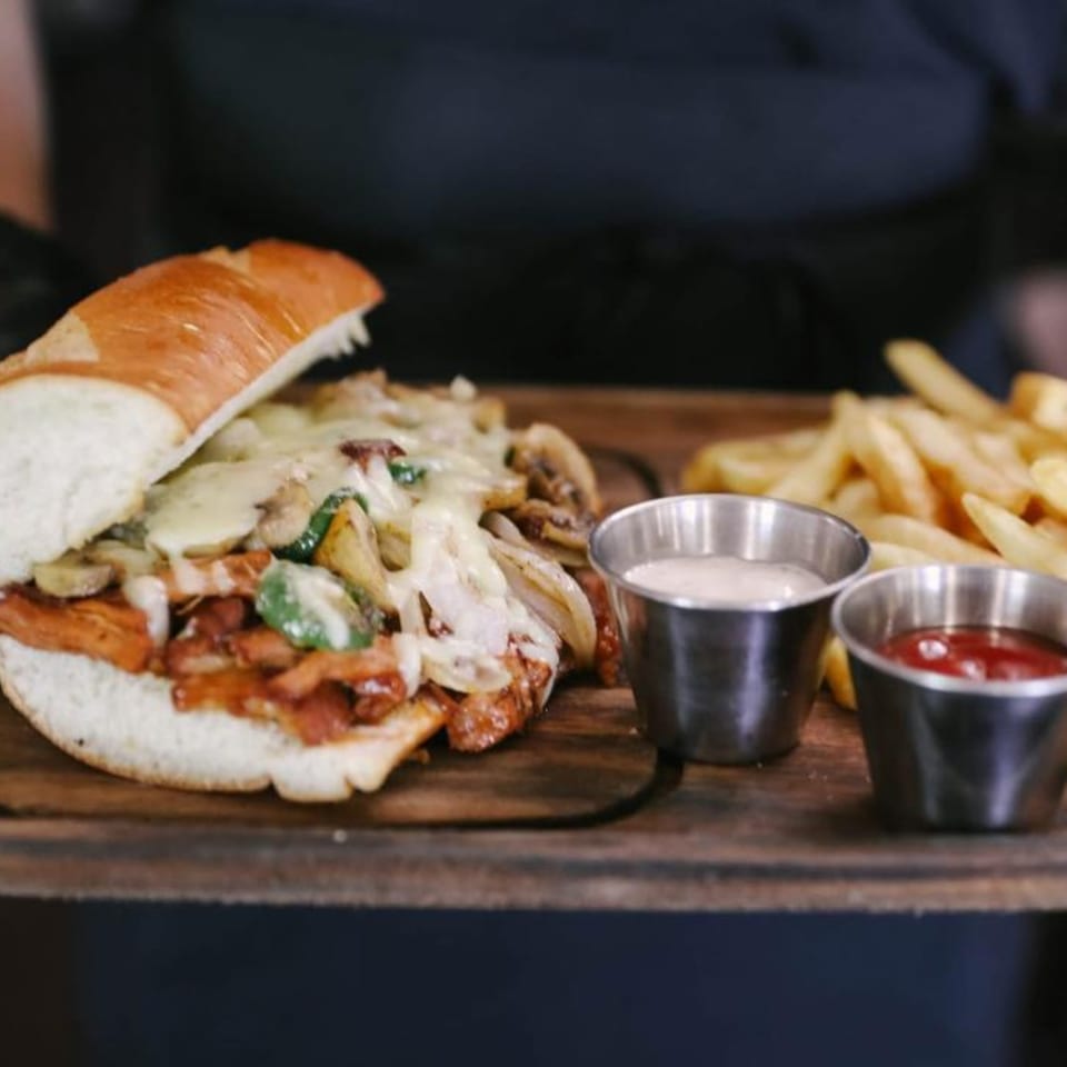 Cheesy steak and mushroom sandwich with fries, ketchup, and ranch on a wooden board.