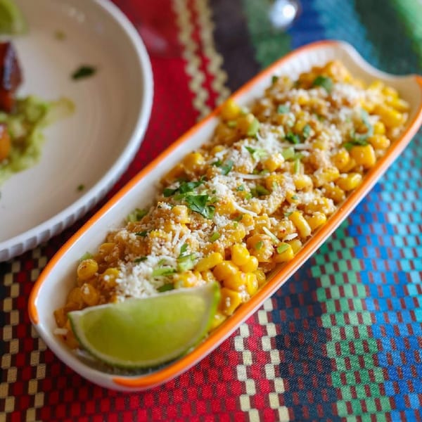 Elote corn in a boat-shaped dish with cheese, cilantro, and a lime wedge on a colorful tablecloth.
