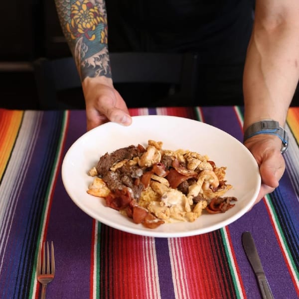 Hands presenting a white plate of mixed meat and eggs on a vibrant striped Mexican serape tablecloth.
