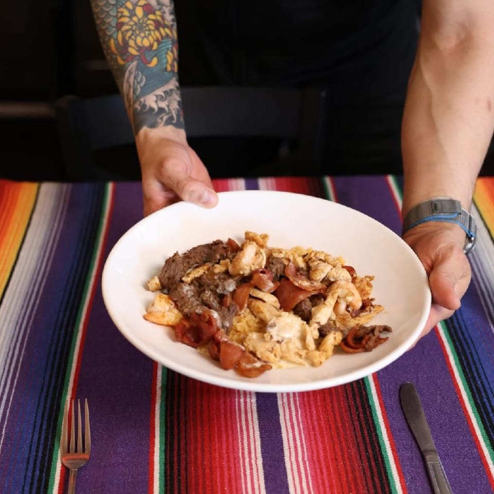 Hands presenting a white plate of mixed meat and eggs on a vibrant striped Mexican serape tablecloth.